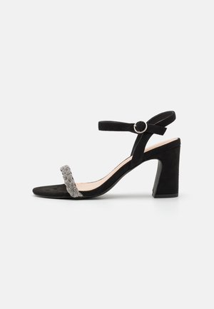 Sort ruskind sandal med høje hæle og ankrem, rund sølvspænde og forrem pyntet med små sølvperler.