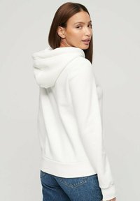 Sudadera blanca con capucha ajustable, textura suave y puños y dobladillo acanalados. Combinada con jeans azules, mostrando un diseño casual y ajustado.