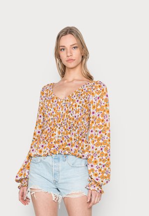 Blusa floral amarela com flores roxas e laranjas, apresenta decote em V, corpo franzido e mangas longas bufantes, combinada com calções de ganga claros.