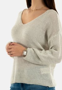 Suéter de punto suelto gris claro con escote en V y mangas largas, combinado con jeans de mezclilla, usado con un reloj dorado y un anillo de plata.