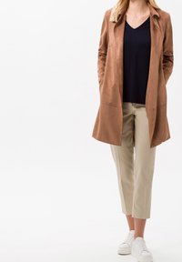 Bruine kunstsuède lange jas, gecombineerd met een marineblauwe V-hals top en beige cropped broek. Witte sneakers maken de outfit compleet.