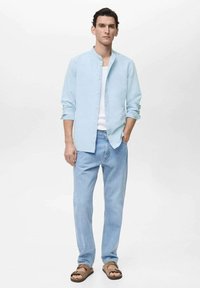 Lichtblauw overhemd met lange mouwen en knoopsluiting, wit T-shirt eronder, gecombineerd met lichte spijkerbroeken en bruine sandalen. Soepele stoftextuur.