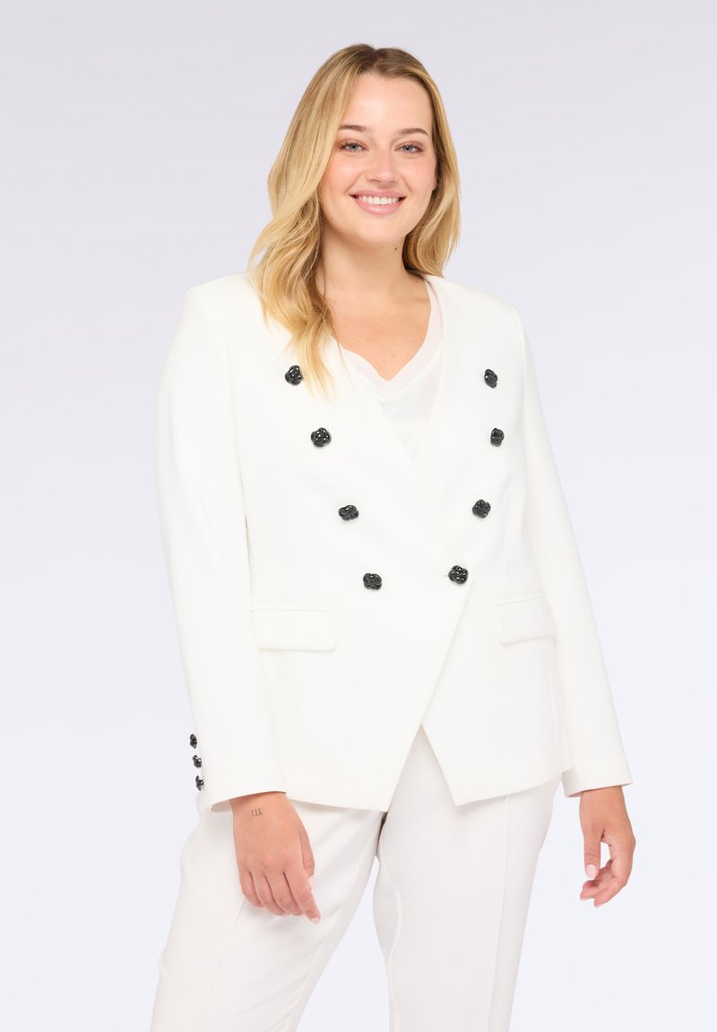 Blazer bianco con bottoni floreali neri, design doppiopetto, rever con notches e tasche laterali, abbinato a una maglietta bianca e pantaloni coordinati.