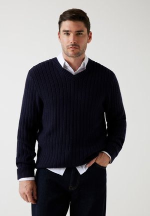 Uomo che indossa un maglione a V a coste blu navy sopra una camicia bianca con colletto, con pantaloni scuri, in piedi contro uno sfondo chiaro e uniforme.