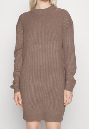 Femme portant une robe pull en maille côtelée à manches longues et longueur genou, couleur taupe marron.