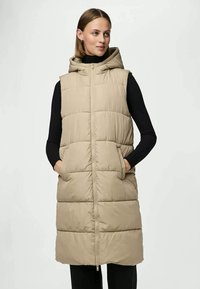 Beige Steppweste mit Kapuze, ausgestattet mit einem durchgehenden Reißverschluss, zwei Seitentaschen und einem durchgängigen Steppmuster. Lange Länge.