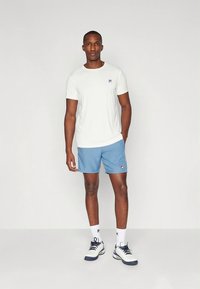 Bela majica z okroglim izrezom, svetlo modre kratke hlače, beli športni čevlji z mornarsko modrimi poudarki, logotip FILA na prsih in hlačah, gladka tkanina.