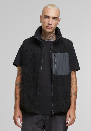 Gilet en polaire noir avec un col montant, fermeture éclair et une poche poitrine gauche en tissu lisse. Surface texturée et coupe décontractée.