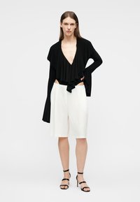 Γυναίκα με μαύρο μακρυμάνικο τοπ με βαθύ V λαιμόκοψη και βολάν, άσπρη βερμούδα μέχρι το γόνατο και μαύρα σανδάλια με ψηλό τακούνι, στέκεται μπροστά σε λευκό φόντο.