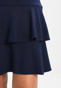 Falda azul marino con dos capas fruncidas, hecha de tela texturizada. El dobladillo es ligeramente curvado, creando una apariencia acampanada en la parte inferior.