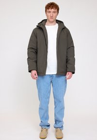 Dark olive green puffer jacket with a hood, zip front, and elastic cuffs, paired with light blue jeans and tan shoes.