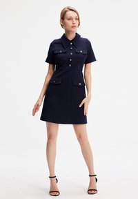 Robe marine avec un col, des manches courtes et des boutons sur le devant. Comprend deux grandes poches poitrine et deux poches taille. Texture lisse.