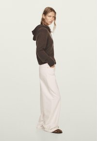 Brown knit hoodie with a front pocket, paired with white wide-leg jeans. Model stands sideways, wearing flat brown sandals.