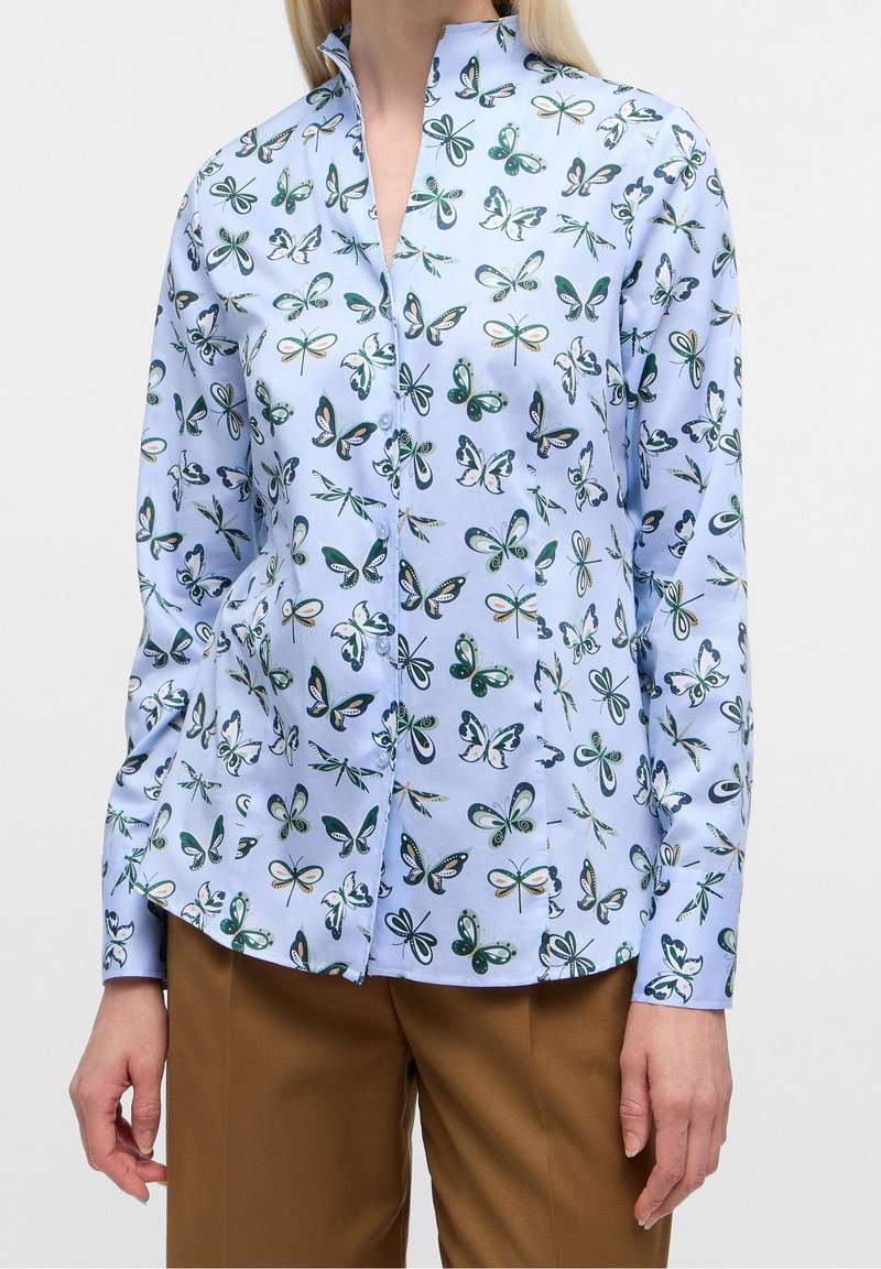 Chemise bleu clair boutonnée avec des imprimés de papillons verts et blancs et de libellules, portée avec un pantalon marron sur une personne debout.