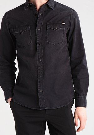 Black long-sleeve button-up shirt with two chest pockets and a small visible brand tag on the left pocket, worn with black pants.