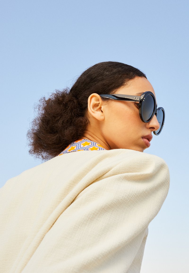 Sorte oversized solbriller med en avrundet design, med gullaccent og en glatt finish. Modellen har på seg en teksturert kremfarget jakke.