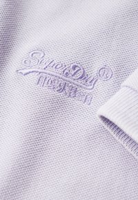 Lichtpaarse katoenen stof met een textuurgebreide structuur. Kenmerkt een geborduurd "SuperDry" logo in een bijpassende kleur. Manchet detail van de mouwen zichtbaar.