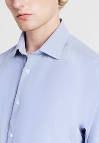 Primo piano di una persona che indossa una camicia a righe sottili di colore blu chiaro con colletto classico e bottoni bianchi.