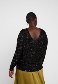 Maglione nero con scollo a V e maniche lunghe, caratterizzato da una maglia strutturata con paillettes argento. Indossato sopra una gonna gialla.