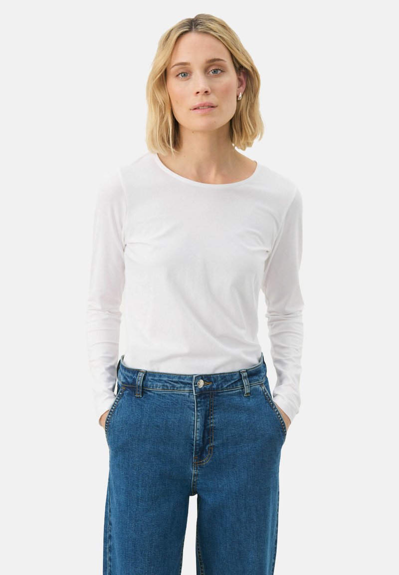Camicetta bianca a maniche lunghe realizzata in tessuto morbido, con scollatura tonda, abbinata a jeans in denim blu larghi, con tasche frontali e chiusura a bottone.