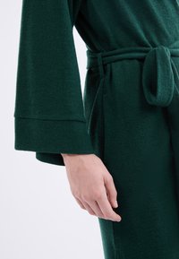 Tmavě zelený, texturovaný hábit s širokými rukávy a odpovídajícím pásem. Má hladkou, měkkou látku a boční kapsy.