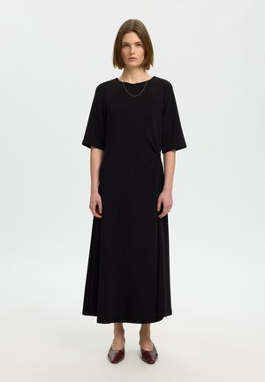 Vrouw met kort bruin haar, gekleed in een lange zwarte jurk met ellebooglange mouwen en donkerrode puntige schoenen, staande tegen een egale witte achtergrond.