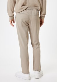 Beige bukser med en afslappet pasform, elastisk talje og baglommer. Materialet virker glat med en subtil glans. Kombinationen med hvide sneakers.