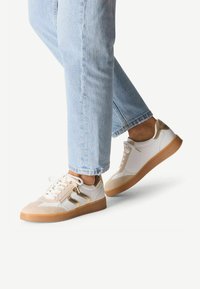 Baskets blanches et beiges avec des accents dorés, présentant un dessus texturé, un laçage et une semelle en gomme. Portées avec un denim bleu clair.