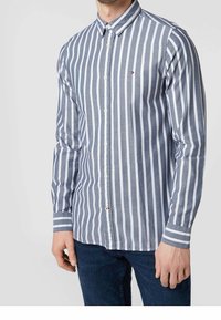 Uomo che indossa una camicia button-up a righe verticali blu e bianche con maniche lunghe e jeans blu scuro.