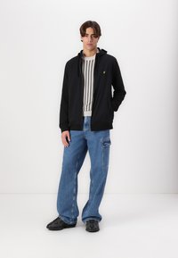 Sudadera negra con cremallera y un pequeño logotipo amarillo, suéter a rayas debajo, pantalones cargo de denim azul sueltos y calzado negro.