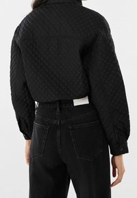 Černá prošívaná bunda s límečkem, texturovaným povrchem a spadlými rameny, kombinovaná s černými džíny s vysokým pasem.