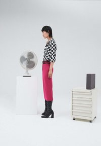 Modella in una camicia bianca e nera a pois, pantaloni rosa brillante e stivali neri fino al ginocchio, in piedi accanto a un ventilatore a piede e a un armadio per documenti.