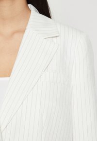 Blazer bianco a righe sottili con revers, taschino sul petto e texture morbida. Le righe sono grigie chiare, aggiungendo un contrasto sottile.