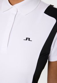 Witte polo met zwarte zijpanelen en een klein zwart logo op de linkerborst, gedragen door een persoon met een gemiddelde huidtint.