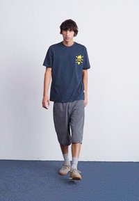 Mørkegrå t-shirt med gul grafik på brystet, parret med lysegrå shorts. Modellen har grå sokker og beige sneakers på.