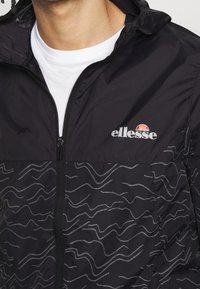 Svart vindjacka med dragkedja. Har ett grått vågmönster och en logotyp på bröstet. Tillverkad av lätt, texturerat material.