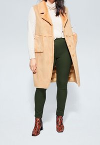 Gilet en fausse fourrure beige avec un col, doté de deux poches avant et de trois boutons, porté par-dessus un pantalon vert et des bottines à imprimé serpent marron.