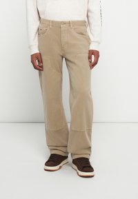 Pantaloni di cotone beige con gambe larghe, tasche frontali e toppe sulle ginocchia, abbinati a sneakers marroni e a una felpa di colore chiaro.