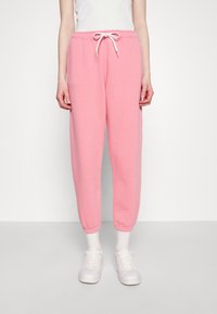 Růžové tepláky s páskem na stažení v pase, zúženými nohavicemi a manžetami na kotnících, vyrobené z měkké tkaniny. Spárované s bílými teniskami a ponožkami.