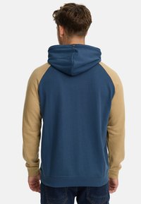 Sudadera con capucha que presenta un cuerpo azul con mangas raglán beige, puños acanalados y una capucha texturizada. Detalle de logotipo sutil en el escote.