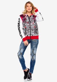 Kapuzensweatshirt in Rot, Schwarz und Weiß; verfügt über ein aufwendiges Grafikdesign, graue und rote Akzente; kombiniert mit verwitterten blauen Jeans und schwarzen Sneakern.