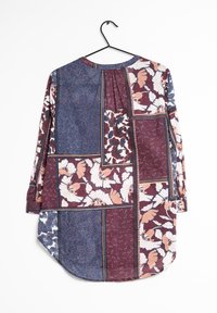 Chemise à manches longues avec motifs floraux patchwork en bleu marine, bordeaux, blanc et pêche, suspendue à un cintre noir sur un fond blanc.