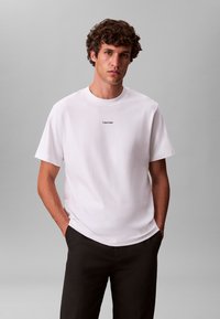 Bijela pamučna majica s okruglim izrezom, kratkim rukavima i malim crnim logotipom "Calvin Klein" na prsima, uparena s tamnim hlačama.