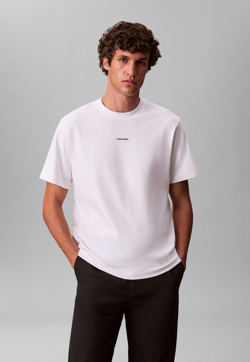 Bijela pamučna majica s okruglim izrezom, kratkim rukavima i malim crnim logotipom "Calvin Klein" na prsima, uparena s tamnim hlačama.