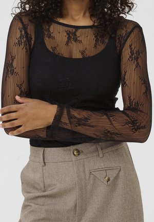 Femme portant un haut en dentelle florale noire transparente sur un caraco noir, associé à un pantalon beige taille haute avec des détails de boutons.