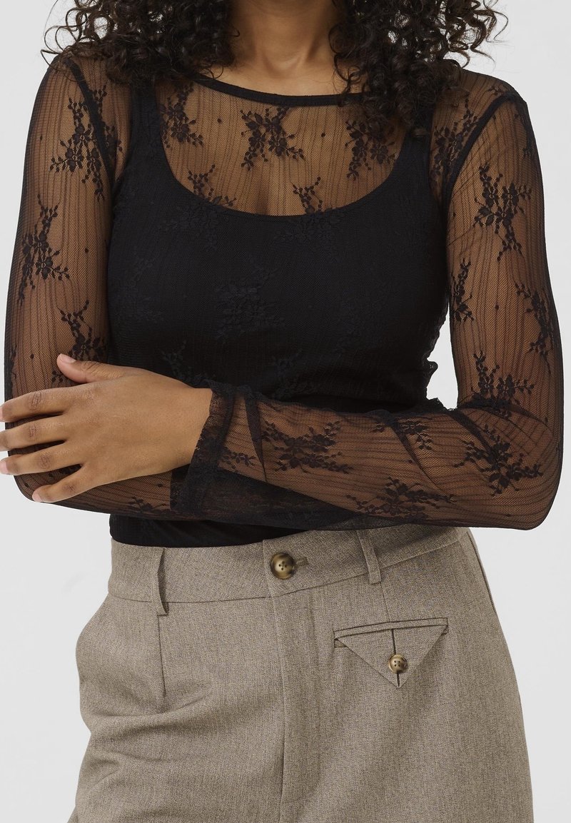 Femme portant un haut en dentelle florale noire transparente sur un caraco noir, associé à un pantalon beige taille haute avec des détails de boutons.