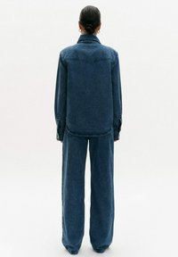 Koszula dżinsowa i spodnie o szerokich nogawkach w ciemnoniebieskim kolorze. Posiada kołnierz, mankiety na guziki i proste cięcia. Gładka tekstura, minimalne detale.