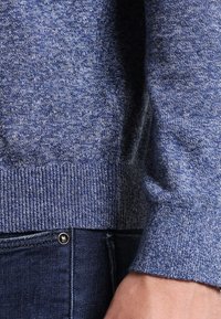 Blauer Strickpullover mit melierter Struktur, ausgestattet mit einem gerippten Bündchen und Saum. Das Kleidungsstück wird über dunkelblauer Denim-Jeans getragen.