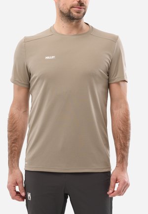 Mann trägt ein beiges kurzärmeliges Sportshirt von Millet und schwarze Shorts von Millet, steht vor einem schlichten weißen Hintergrund.