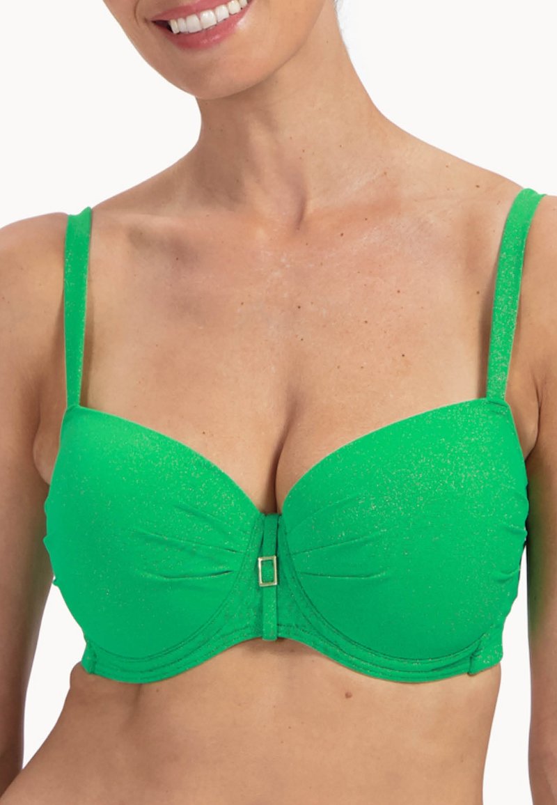 Mujer con un top de bikini verde brillante con tirantes delgados y un pequeño broche central, sonriendo con los dientes visibles sobre un fondo blanco.